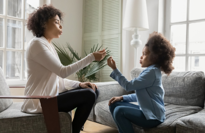 An adult black woman works with a young black girl. They sit together in a living room on couches.